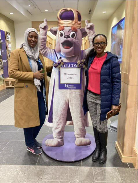 Two women smile and pose for a picture inside while standing beside a cutout of the JMU mascot "the Duke Dog".