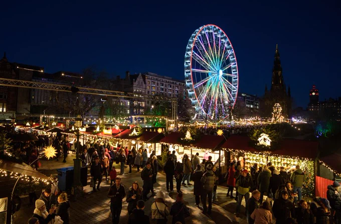 Night shot with crowd of people in front of market stalls in the foreground and big wheel in the background. 
