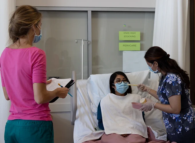 A nursing assistant student is holding a toothbrush for another student who is acting as a patient in a bed. An instructor is supervising. 
