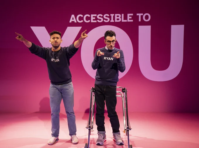 Dickie Hearts and Ryan J. Haddad stand on a stage with pink flooring and a pink wall behind them with the words “ACCESSIBLE TO YOU” written in white. The word “YOU” is much larger than the rest of the text. Ryan is a Lebanese American man with short black hair, brown eyes, and round, tortoiseshell glasses. Ryan wears a navy blue long-sleeve sweatshirt with the word “RYAN” printed in white across his chest and a white button-down shirt underneath it. His walker stands in front of him. He wears blue jeans and grey and white Nike sneakers with red and black soles. Dickie Hearts has olive skin and dark chocolate-colored curly hair. He also wears a navy blue long-sleeve sweatshirt with the words “RYAN” printed in white across his chest. He wears light blue jeans and white sneakers with rainbow soles. Both actors point to the audience with serious faces. Dickie’s arms are outstretched, and Ryan’s arms are bent. They both point forwards with their index fingers.
