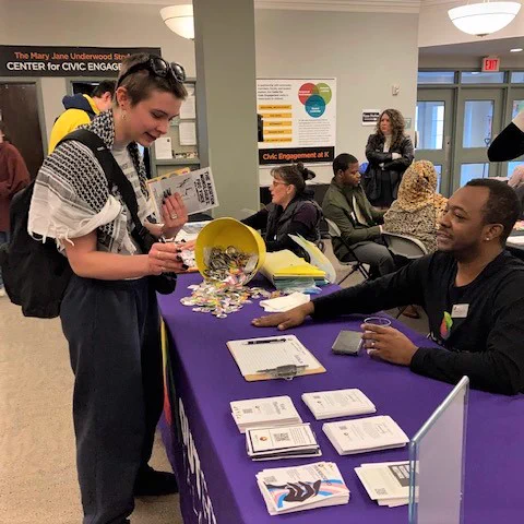 Student speaks with community leader at OutFront Kalamazoo at their table with purple tablecloth and many flyers and buttons spread out.