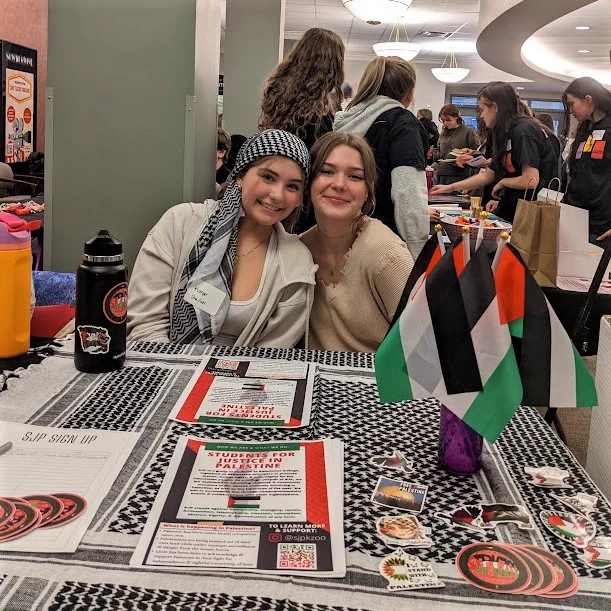 Two students tabling for the student organization Students for Justice in Palestine smile and pose for a picture. Their table has Palestinian flags, flyers for the club, and stickers to take.