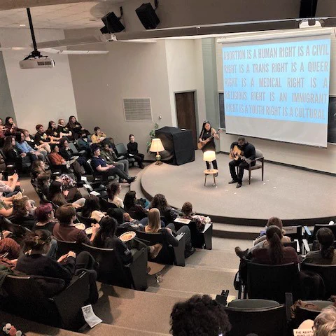View of small auditorium full of people watching two performers on stage. One standing and the other sitting, both playing string instruments and singing. 