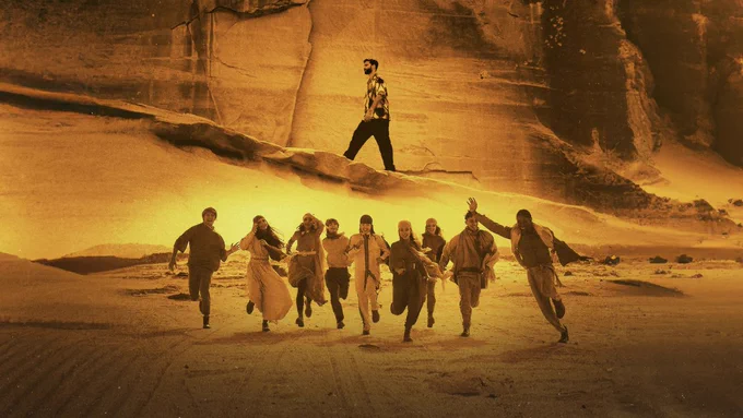 A sepia image of the members of Now United, all wearing desert garb, running through the desert, with R3HAB, wearing black pants and a button-down shirt, walking up a mountain in the background.