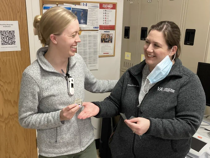 Dr. Sylvia Keiser receives a silver coin from Dr. Michelle Brooks in a work area. Both physicians are smiling.