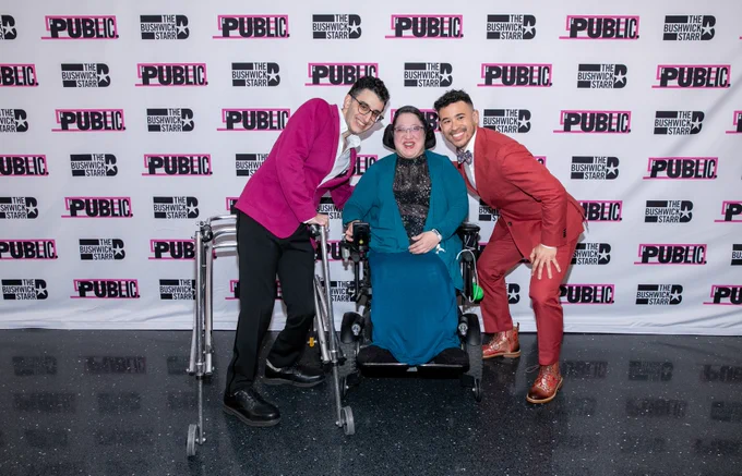 Ryan J. Haddad, Alejandra Ospina, and Dickie Hearts stand in front of a white Step & Repeat, posing for a photograph. Ryan wears a magenta suit jacket, a white button-down, black pants, and black dress shoes. His walker stands in front of him. Ryan is a Lebanese American man with short black hair, brown eyes, and round, tortoiseshell glasses. Dickie wears a light red suit, a floral bowtie, a white button-down, and brown and red dress shoes. Dickie Hearts has olive skin and dark chocolate-colored curly hair. Alejandra Ospina wears a light blue sweater, a black blouse with white dots on it, and a blue skirt. Alejandra, a light-skinned Latina with short dark hair that is slicked back, sits in her motorized wheelchair. The Step & Repeat behind them says The Public with pink lines and a box behind it and The Bushwick Starr with a star next to it.