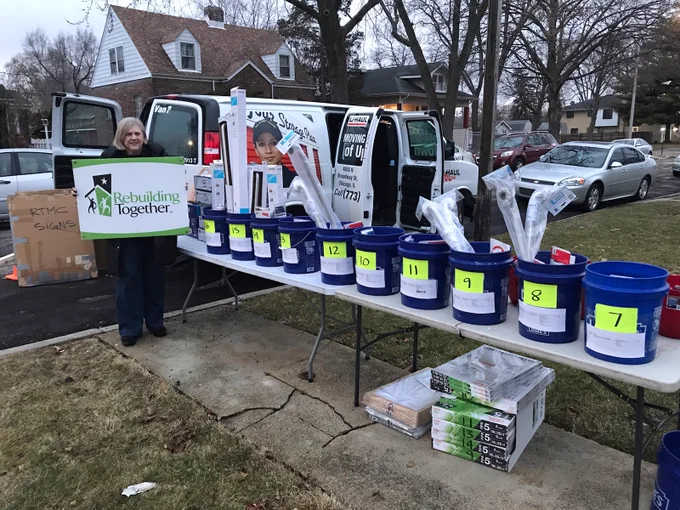 Rebuilding Together Metro Chicago volunteer next to tables with blue buckets on them full of materials to make home repairs and modifications. 