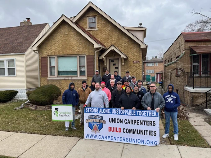 A group of about 20 Local 10 volunteer carpenters standing in front of one of the homes in which they are going to be making some modifications. Three of the volunteers in front are holding the Mid America Carpenters Regional Council Sign that reads "Strong Alone. Unstoppable Together. Union Carpenters Build Communities."