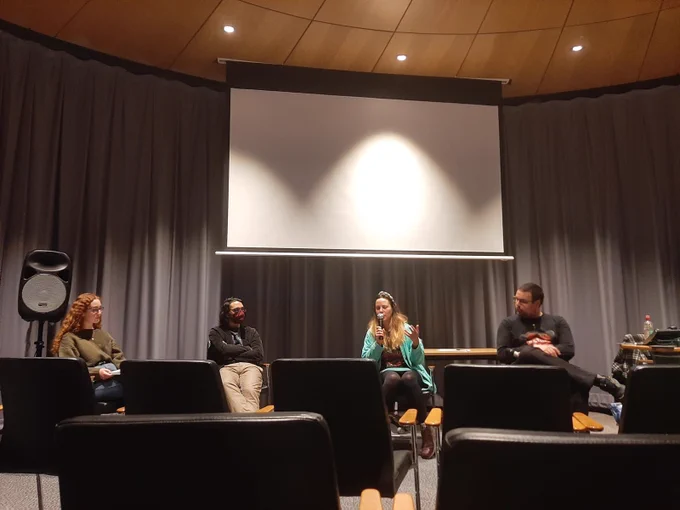 Four people sit on a panel discussion. Left to right the panelists pictured are: a woman with curly red hair, a main with dark hair and a red covid-19 mask, a woman with long brown and blonde hair speaking into a microphone and a man with short brown hair.