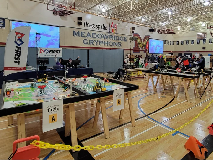 FIRST LEGO League Challenge Event in a Gymnasium with LEGO kits in wooden tables I'm the foreground & students at worktables in the background 