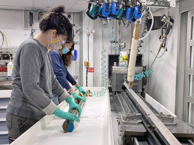 Two scientists (me and a friend, both wearing masks, safety glasses and gloves) holding a freshly split core-section of deep-sea sediment. We are in the core splitting room in the Core Lab of the JOIDES Resolution, and a variety of machinery is visible on either side of us. Photo credit: JRSO-IODP.