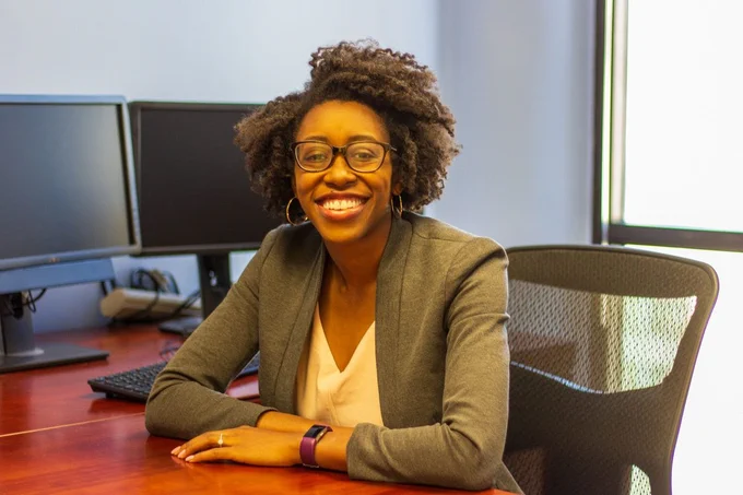 Dr. Sierra Carter sits at a wooden desk with her arms folded and resting on the desk. She wears a white blouse and gray blazer with hoop earrings and black-frame glasses. She smiles at the camera, with two computer monitors and a bright window behind her.
