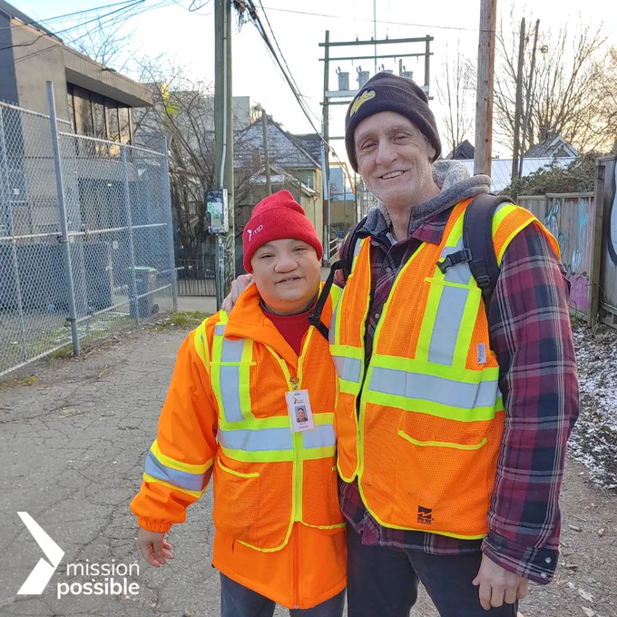 Ben and Real stand next to each other in an alley way smiling. They are wearing orange safety vests and beanies. Ben is wearing a bright orange jacket underneath and Real is wearing a plaid one. In the bottom left corner is Mission Possible's logo in white.
