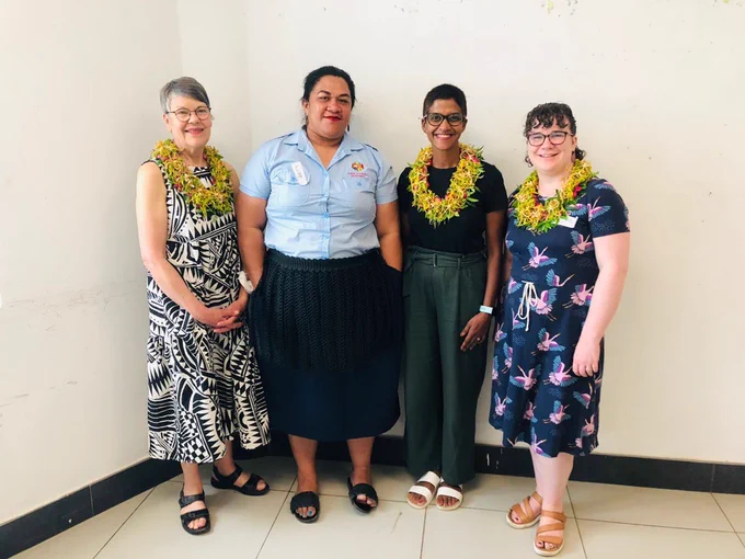 Four women are smiling for a photo. Two woman are white and two are brown. Some are wearing a flower lei. They look happy to be working together. 