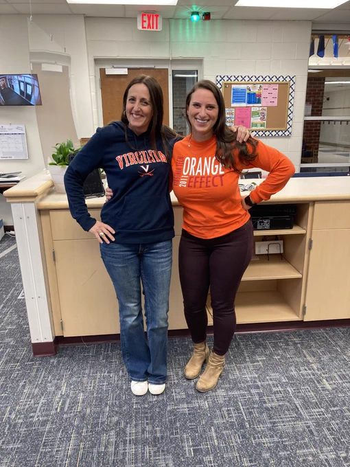 a picture of two women in an office, one wearing a navy blue Virginia (UVA) shirt, and one wearing an orange Virginia Tech shirt.