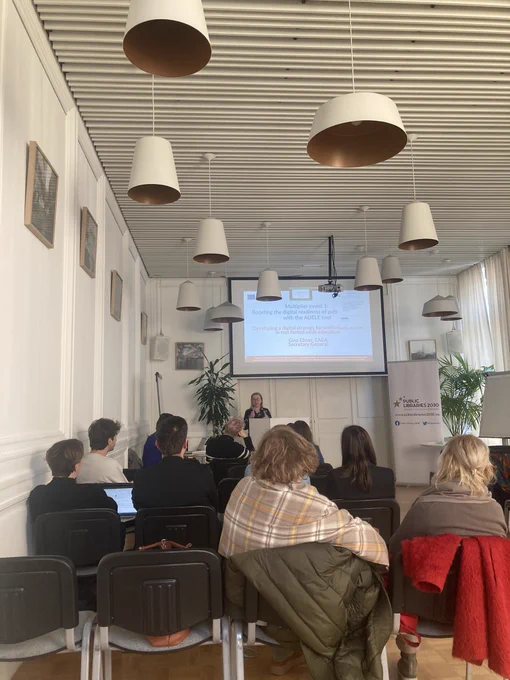 Photo of people sitting in a room. One person at the front speaking in front of a presentation