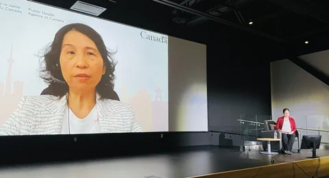 Dr Tam, wearing a white jacket with black cursive writing, in projected on a large screen on the left. Sitting on a chair at right in a red jacket is interviewer Michelle Campbell Mekarski