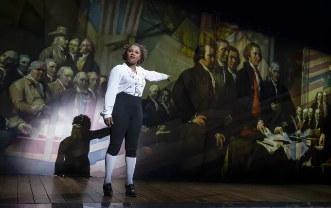 Gisela Adisa, as 'John Adams', points at a curtain projection of the Historical Painting of the Founding Fathers in Congress at the top of the show. 