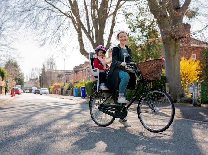 A woman on her bike. She has her child on a child seat behind her and she has a basket on the front
