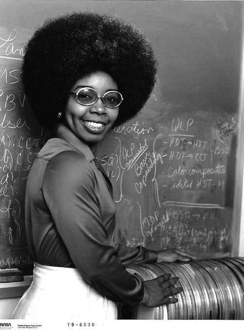 A black and white image of Dr. Valerie Thomas in 1979 standing with a stack of early Landsat Computer Compatible Tapes (CCTs). Behind Dr. Thomas is a blackboard covered in computations. She smiles at the camera wearing round glasses, her hair in an afro, she is wearing a blouse with a white skirt. Credit: NASA