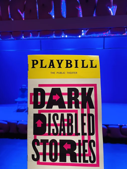 Image description: 
Photo of the Dark Disabled Stories playbill cover held up in the foreground, with the set glowing in a blue light in the background of the shot. The playbill cover is in black text on top of a background pink outline design that boldly and largely reads: "Dark Disabled Stories", with a yellow “Playbill" logo strip across at the top.