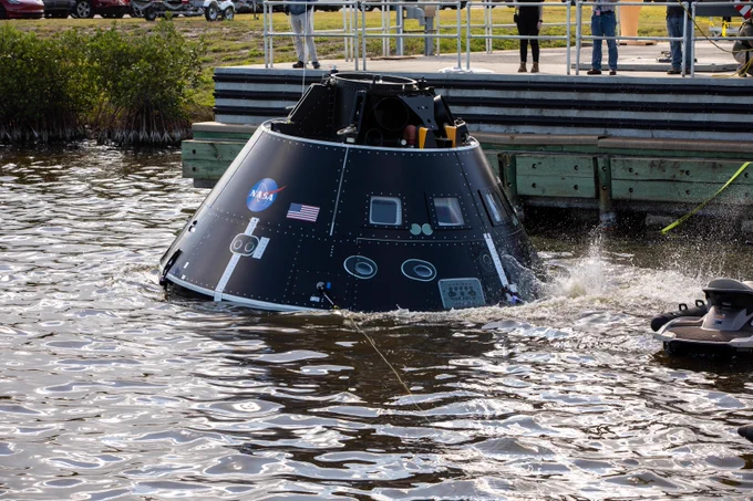 The black and silver CMTA lands in the test basin, causing the steel-black water to churn up. A jet ski is visible on the right, with onlookers behind the mock-up module in the background.