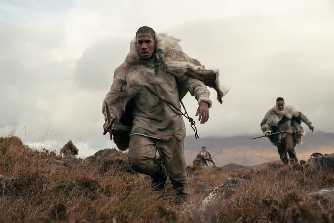 Still from THE ORIGIN of a group of Stone Age wanderers running on the hills in the Scottish Highlands. They wear fur pelts and wield spears.
