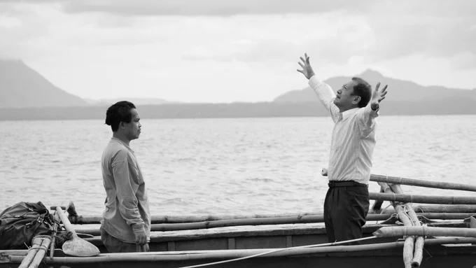 Still from WHEN THE WAVES ARE GONE of two men stood on a small boat in the water with mountainous landscapes behind them. One of the men looks up with his arms outstretched. 