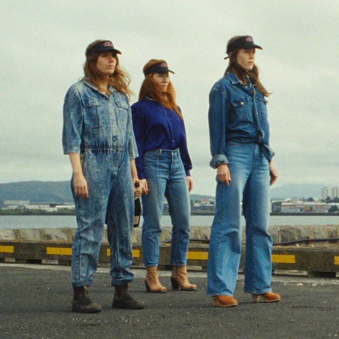 Still from BAND of three women with long hair and mainly-denim outfits with caps staring out to sea. 