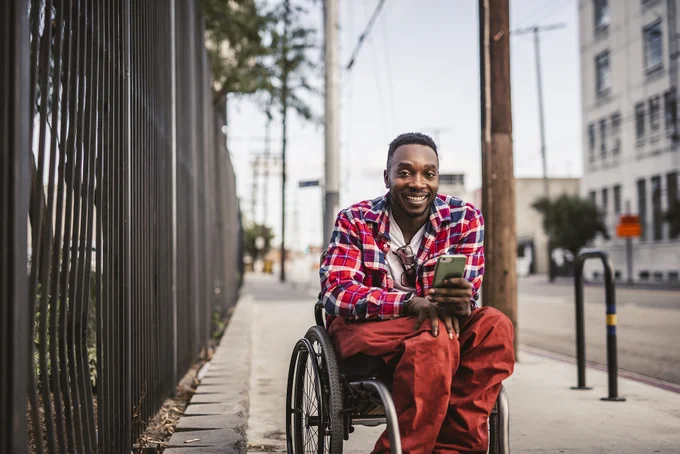 A person smiles, looking at the camera. They are in a wheelchair and holding a mobile phone. They are outside on the pavement. 