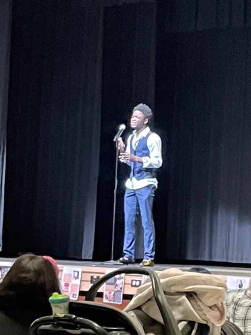 A Black male student performing an original song in front of a microphone on a stage.