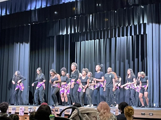 Students wearing all black with purple African fabric wraps on their waists on a stage performing a dance