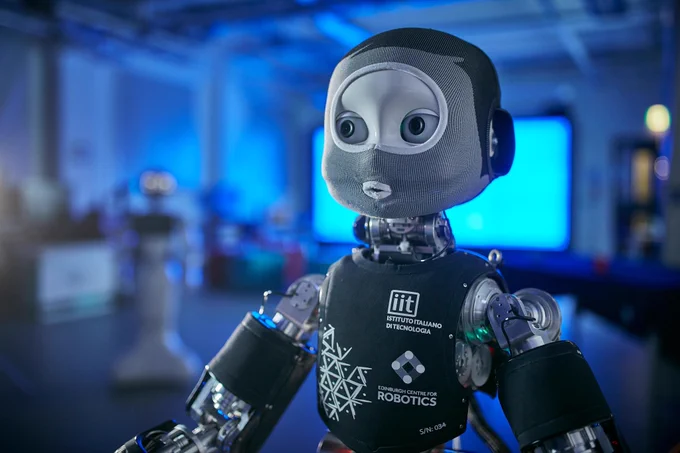 An anthropomorphic robot wearing a mesh suit against a blurred background showing a blue lit laboratory. 
