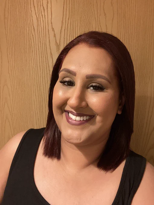 Photo of Simran standing against a brown wooden door. She is wearing a black vest top, has her hair down, styled straight with light makeup on and is smiling big.