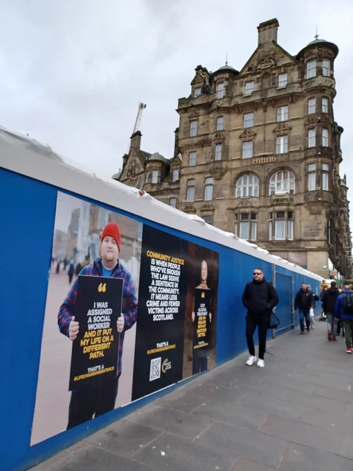 Two large posters with people holding billboards and one with text only on North Bridge in Edinburgh