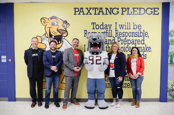 Five people and a bulldog mascot pose in front of a wall featuring the Paxtang Pledge in an elementary school