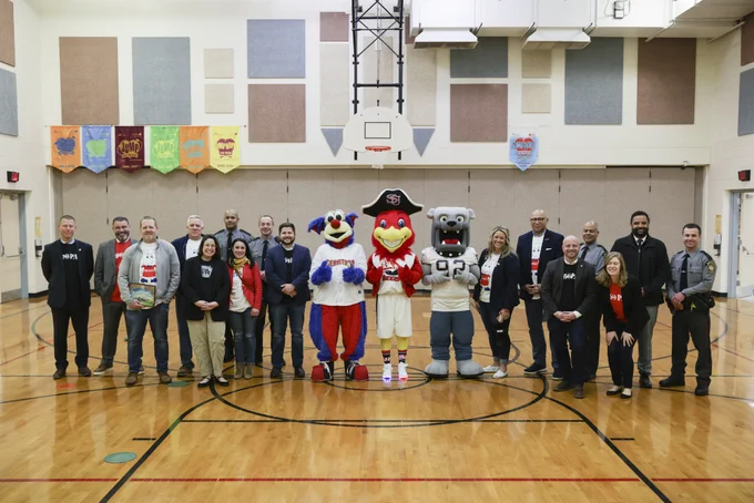 Sixteen people and three mascots pose for a picture in an elementary school gymnasium