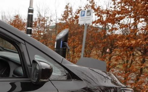 Photograph of the bonnet of a black car with an electric vehicle charger in the background, which is plugged into the car. A sign with the parking symbol is in the background with bushes behind that