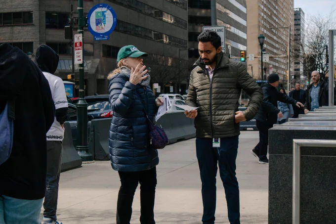 One person in a blue coat gestures with their hand as they talk and another person in a green coat looks down at a paper flyer as they listen.