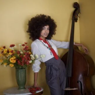  Esperanza Spalding leans against a wall with a vase of flowers to her right and cello to her left.