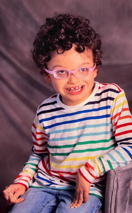 A school photo of an 8-year-old boy. He has curly brown hair and is wearing glasses and a rainbow-striped shirt.