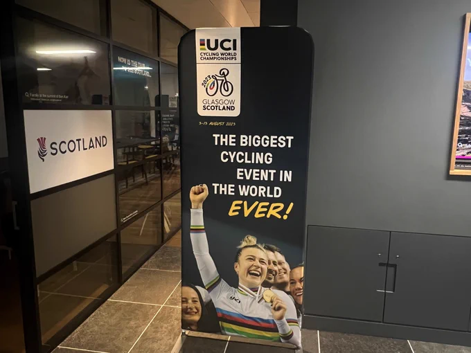 A photograph of a pop-up banner featuring a group cheering. Text 'The biggest cycling event in the world ever!' 