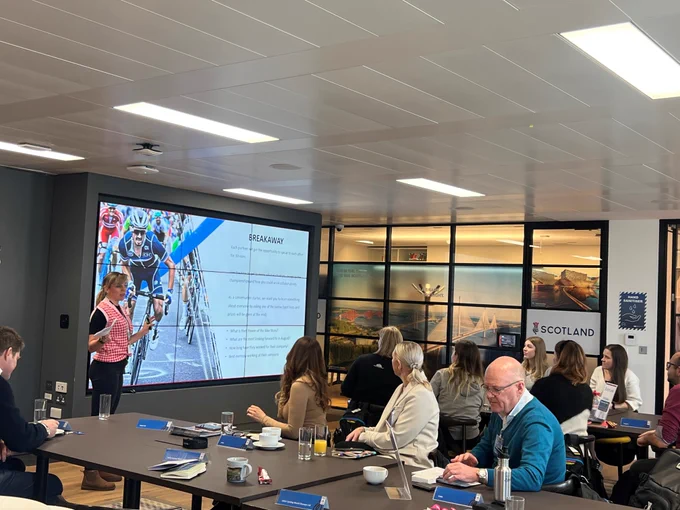 An image of someone delivering a presentation to a group of people sat around tables