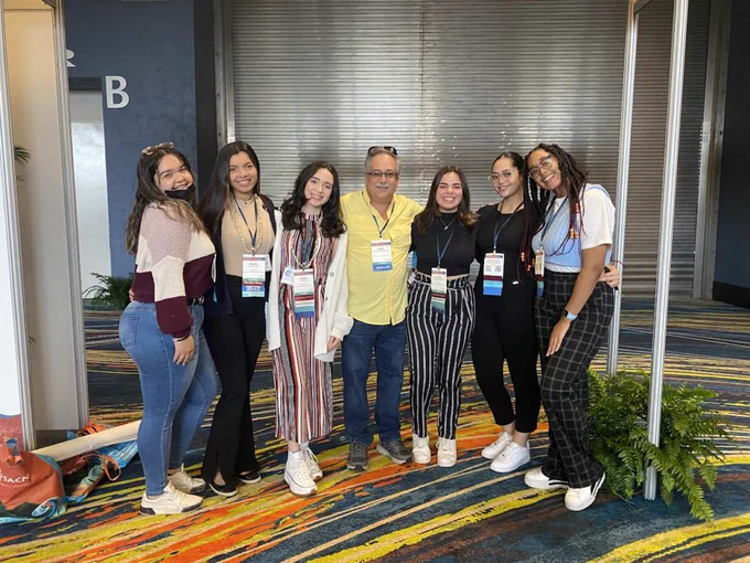 Seven people stand in a row smiling. They are wearing conference badges and stand on a colorful, striped carpet with a metal wall behind them.