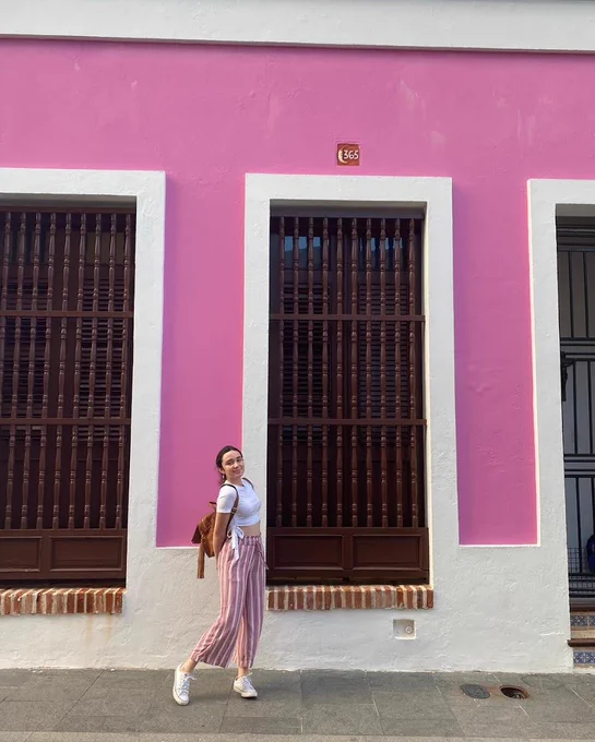 A woman wearing pink and white striped pants, a white cropped t-shirt, white sneakers, and a brown backpack stands in front of a pink wall. The wall has two large windows with wooden bars and white trim.