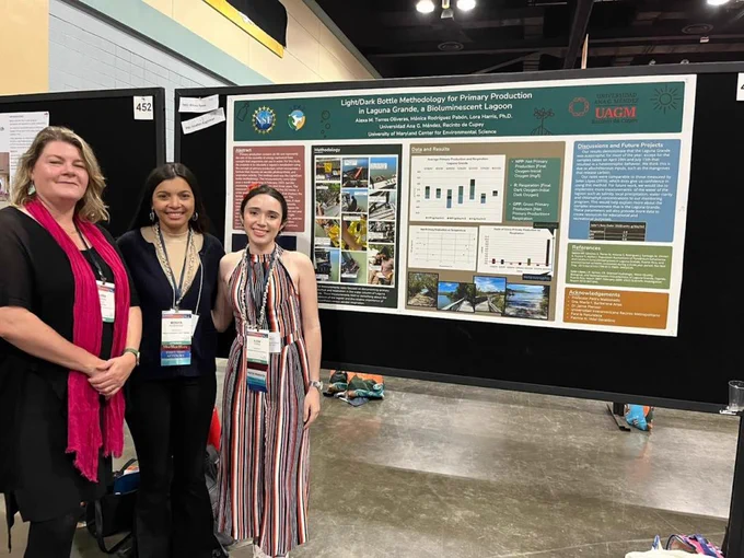 Three people stand smiling in front of a research poster. They are wearing conference badges.