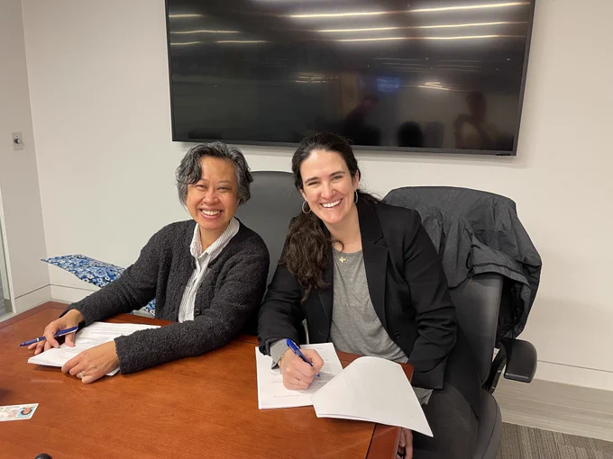 Two women sitting at a table smiling. They have documents on the table in front of them. 