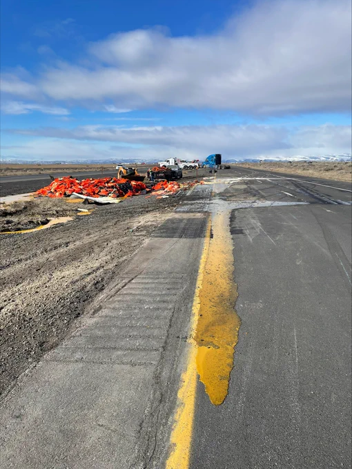 Egg yoke runs along the yellow line of the left lane of highway, to the left in the median there is a pile of orange crates. There are several vehicles in the background including two semis and pick ups with trailers.