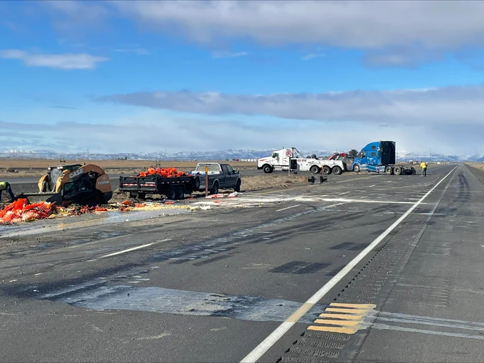 
Egg yoke runs across the highway, to the left in the median there is a pile of orange crates being cleared by a forklift. A large semi tow truck is loaded with the crashed semi in th ebackground.