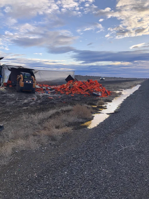 A forklift is clearing broken piles of crates from a shattered and overturned semi trailer on the grass median of the highway. A large pile of crates is beside the broken trailer and a substantial puddle of yellow egg yolk is polling against the highway shoulder.